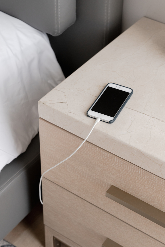 A smartphone is placed on a beige nightstand, connected to a white charging cable. The bed is partially visible on the left side.