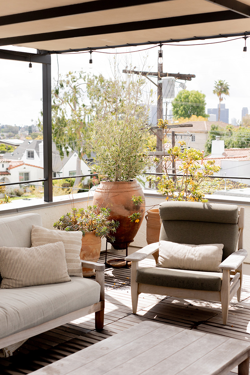 Rooftop patio with beige furniture, large potted plants, and a view of distant buildings under a cloudy sky.