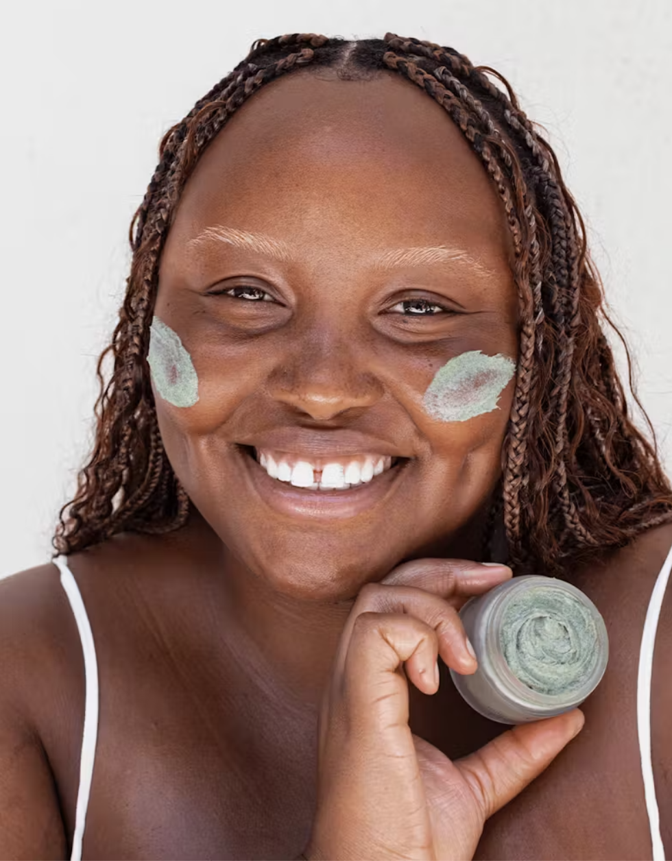 A person with braided hair and a bright smile holds a jar of face cream, with green cream applied to both cheeks. They are wearing a white top against a plain background.