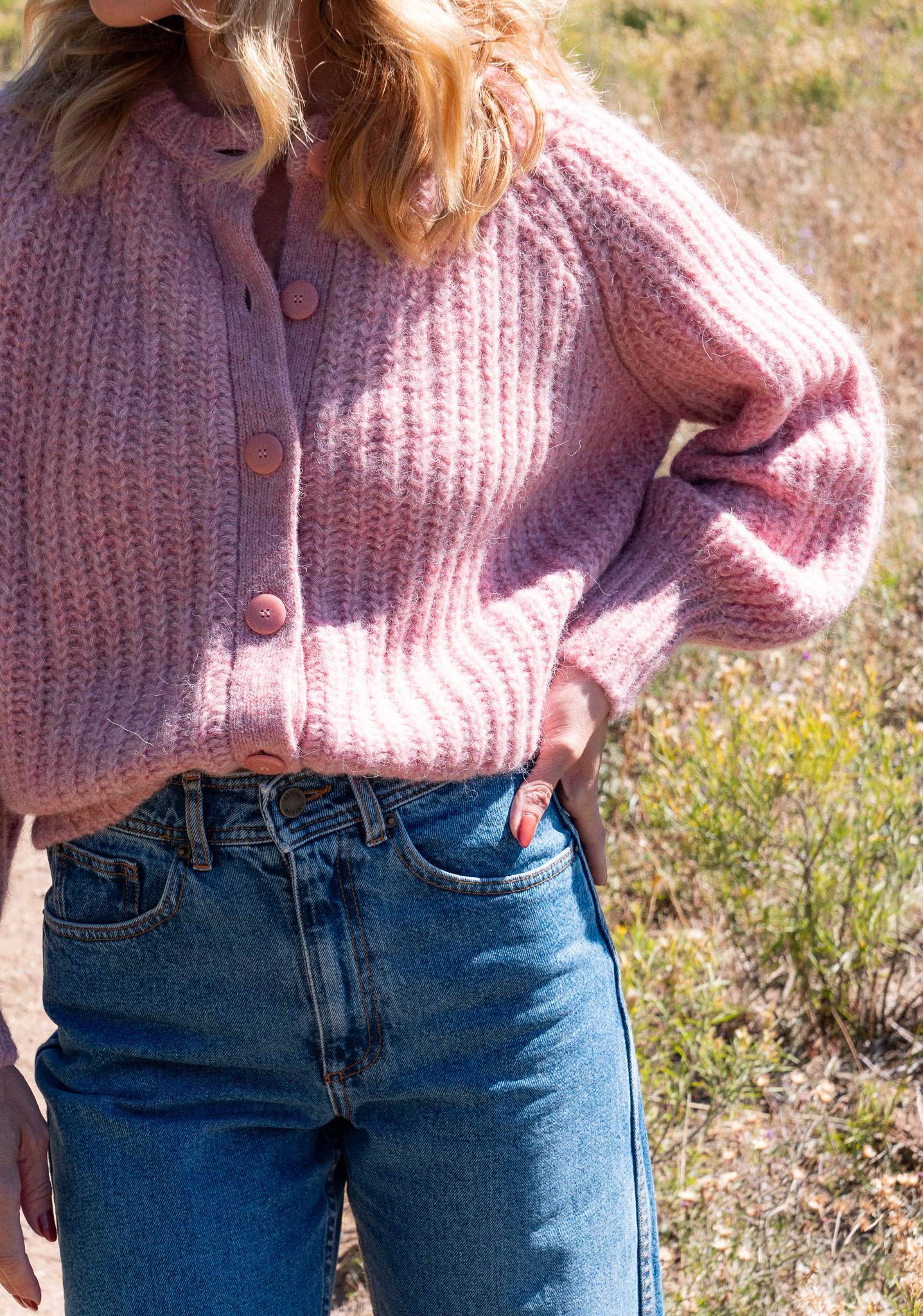 Person wearing a pink knit sweater with large buttons and blue jeans, standing outdoors with hand in pocket.