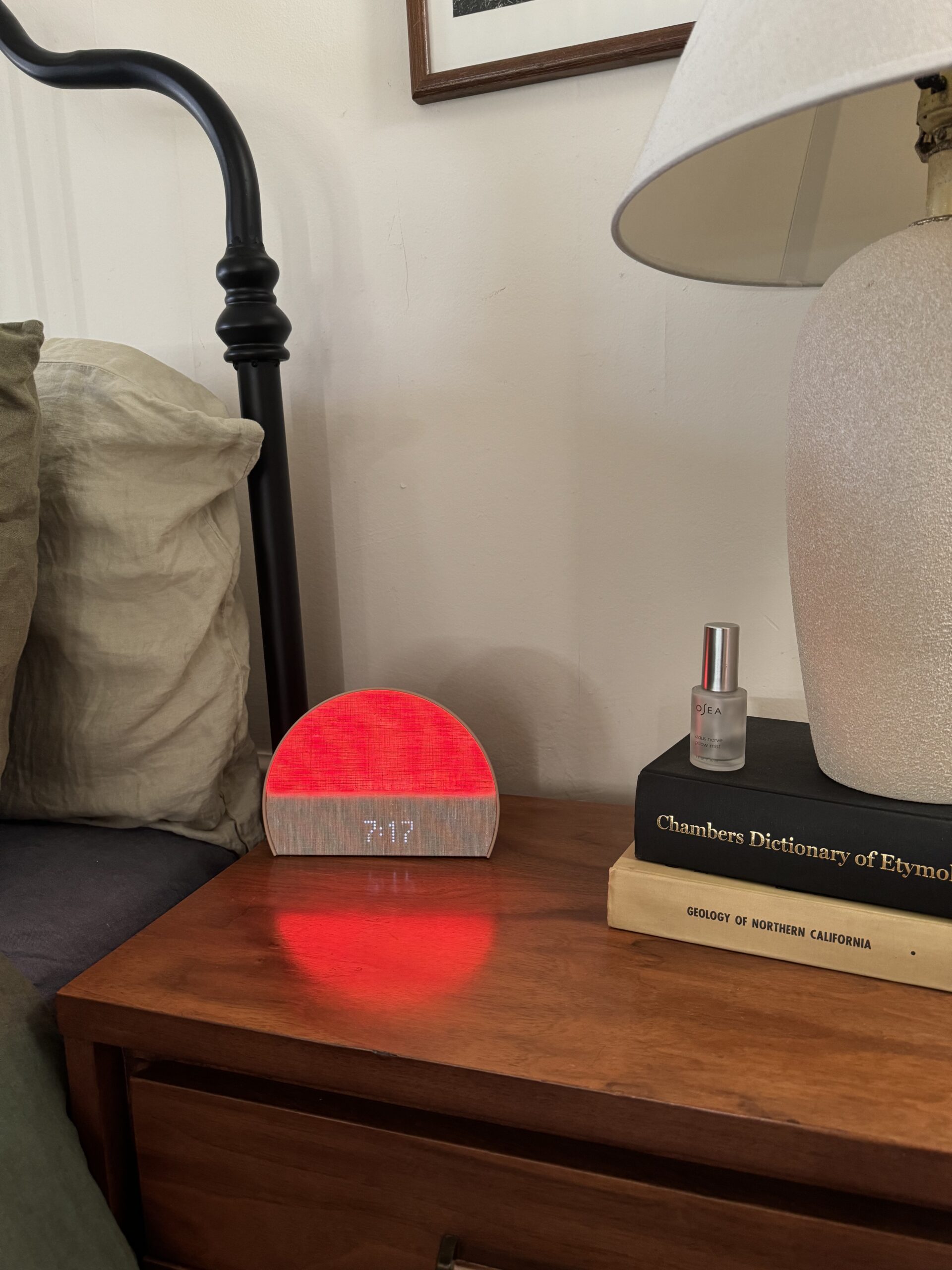 A wooden nightstand with a digital alarm clock displaying 7:17, a lamp, a small spray bottle, and a stack of books, including "Chambers Dictionary of Etymology.