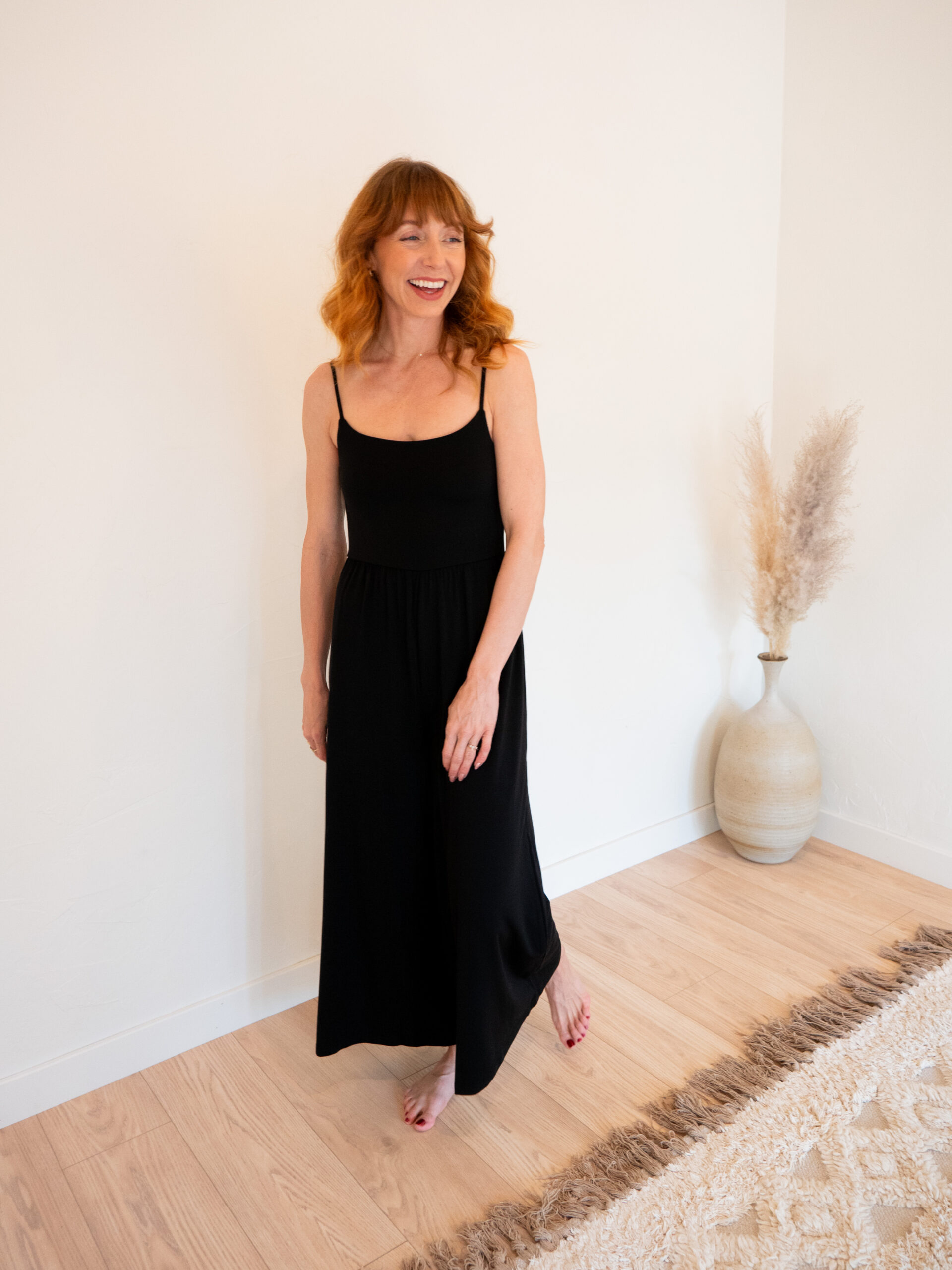 A person with red hair stands barefoot on a wooden floor, wearing a sleeveless black dress next to a tall vase with pampas grass.