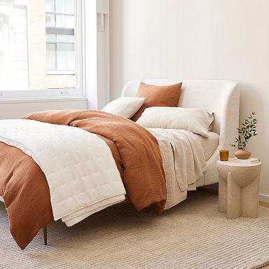 A neatly made bed with white and rust-colored bedding, positioned near a window. A small side table holds a plant and a glass. The room has a light, neutral color scheme.