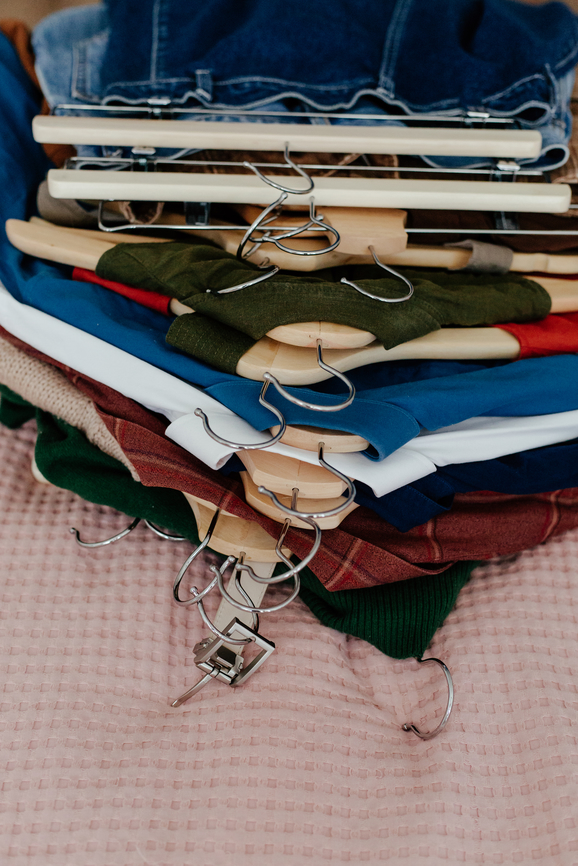 A stack of various colored clothes neatly hung on wooden and metal hangers, placed on a pink textured surface.