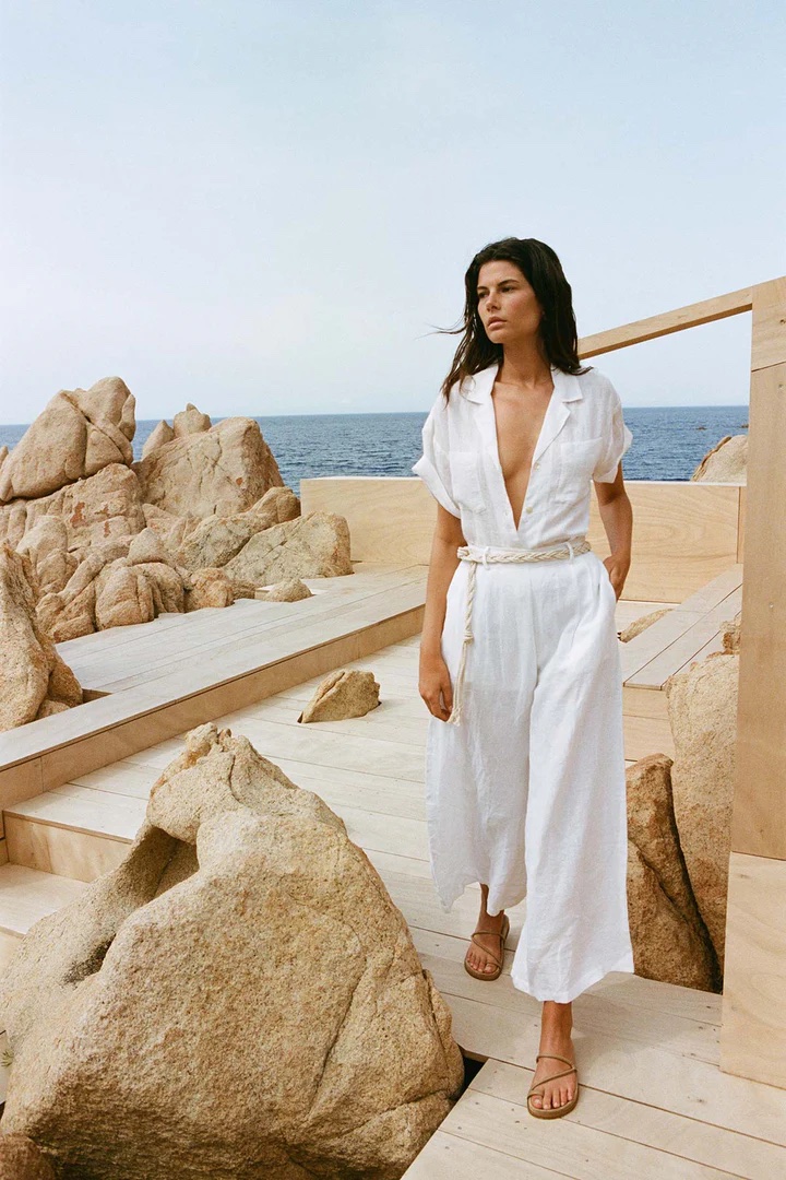 A woman in a white dress stands on a wooden deck near rocky terrain with the ocean in the background.